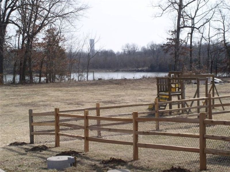 Wooden fence encloses a playground; beyond, a lake and treeline, a water tower visible.