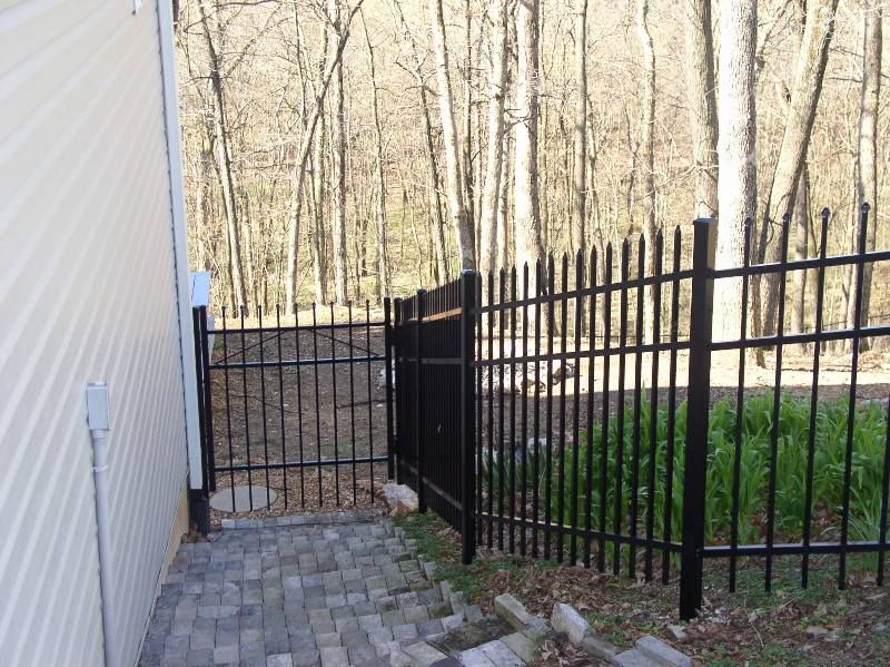 Black metal fence and gate alongside a brick path and a beige building, leading into a wooded area.