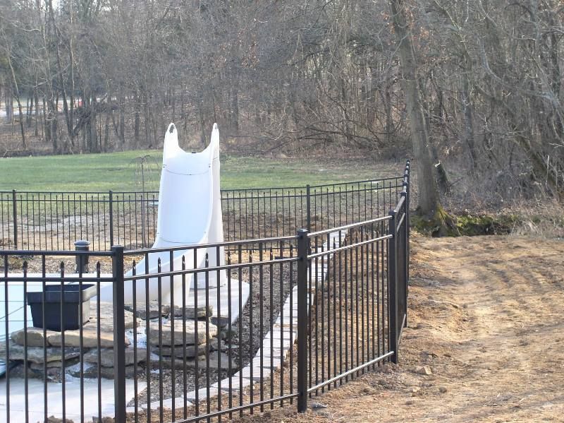 Black metal fence surrounds a pool with a white slide in a yard with trees.