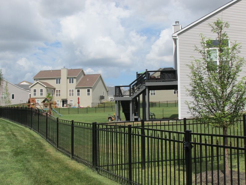 Black fence curves around a grassy backyard, with houses in the distance under a cloudy sky.