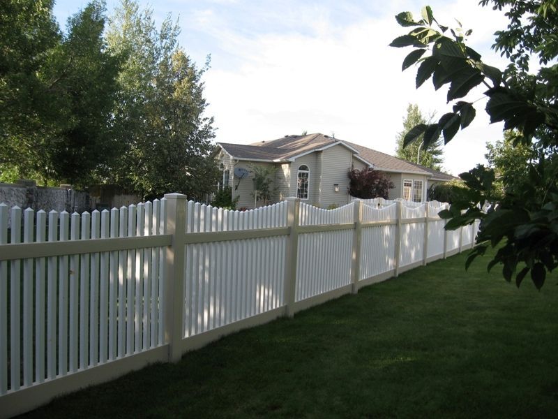White picket fence surrounds a house with a green lawn and trees in the background.