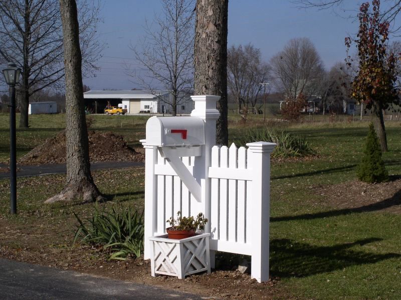 White mailbox with decorative picket fence, small potted flowers, roadside setting.