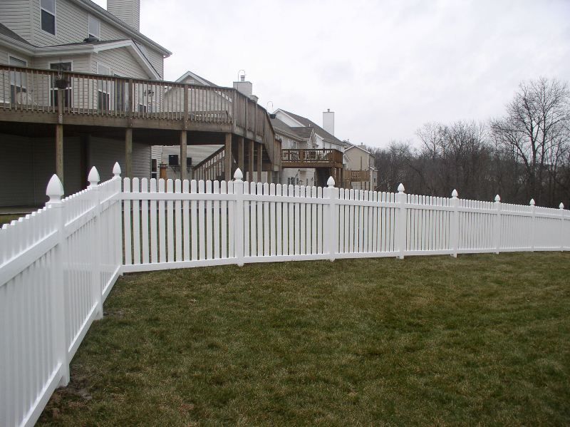 White picket fence surrounding a grassy yard, a house with a deck in the background.