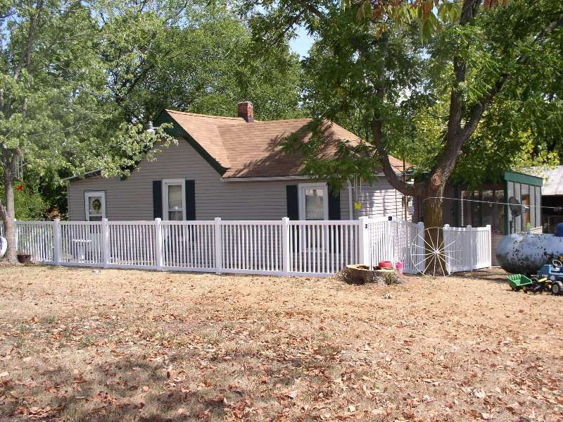 Small, weathered house with white picket fence, surrounded by trees and dry leaves.