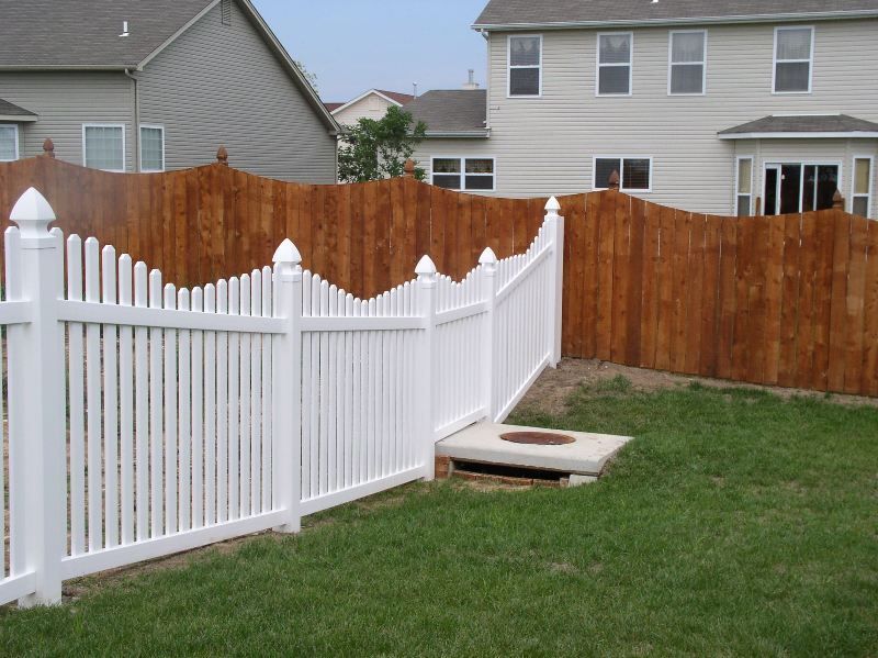 White picket fence and wood fence enclosing a green lawn in a backyard.