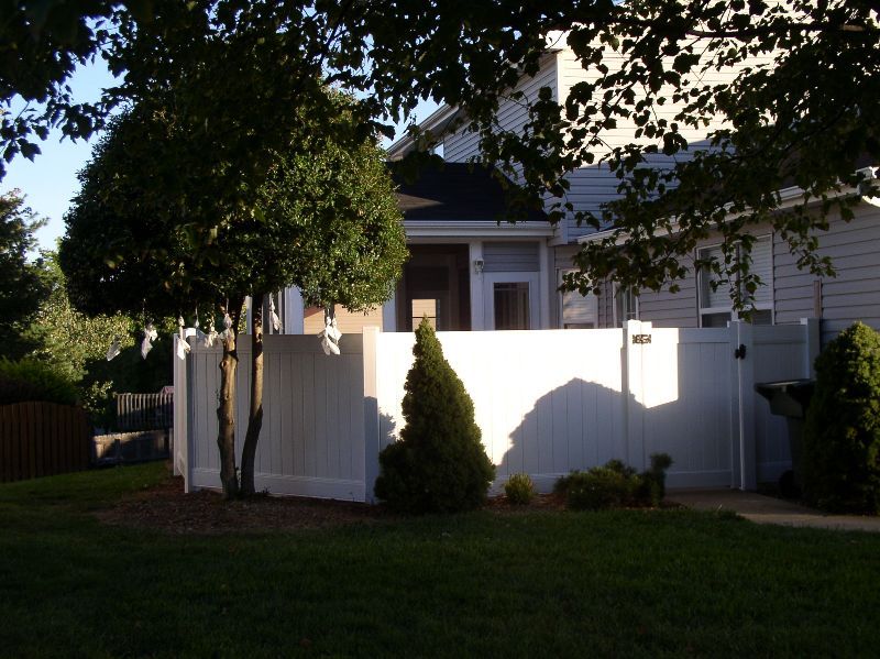 White privacy fence surrounds a backyard with a house visible in the background; trees cast shadows on grass.