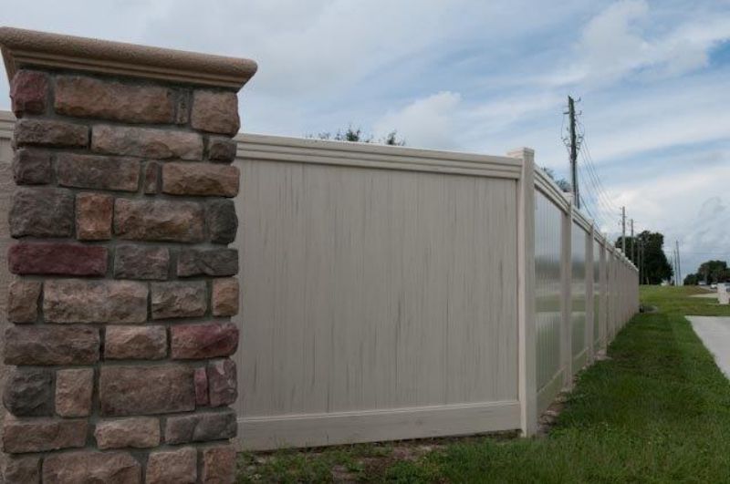 Stone column next to a beige privacy fence along a grassy curb and road under a partly cloudy sky.