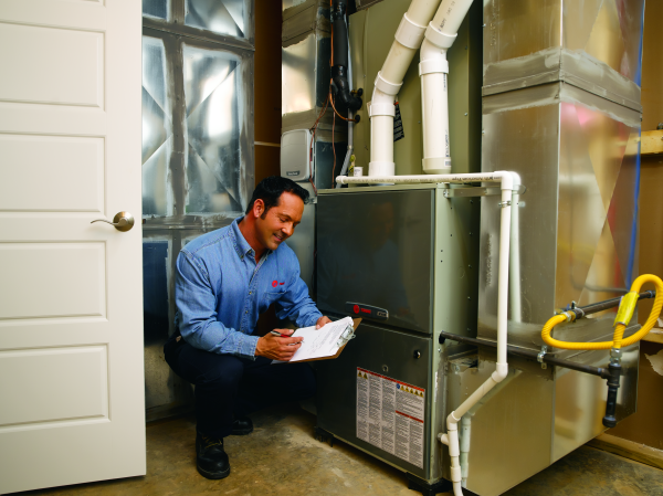 A man is kneeling down in front of a furnace and writing on a clipboard.
