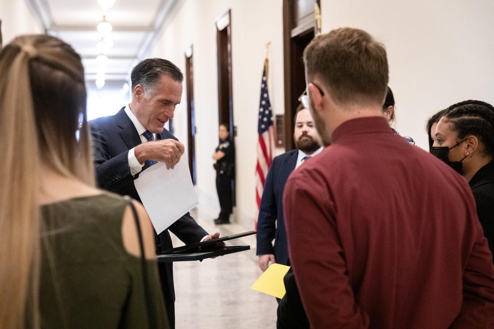 A man in a suit and tie is talking to a group of people in a hallway