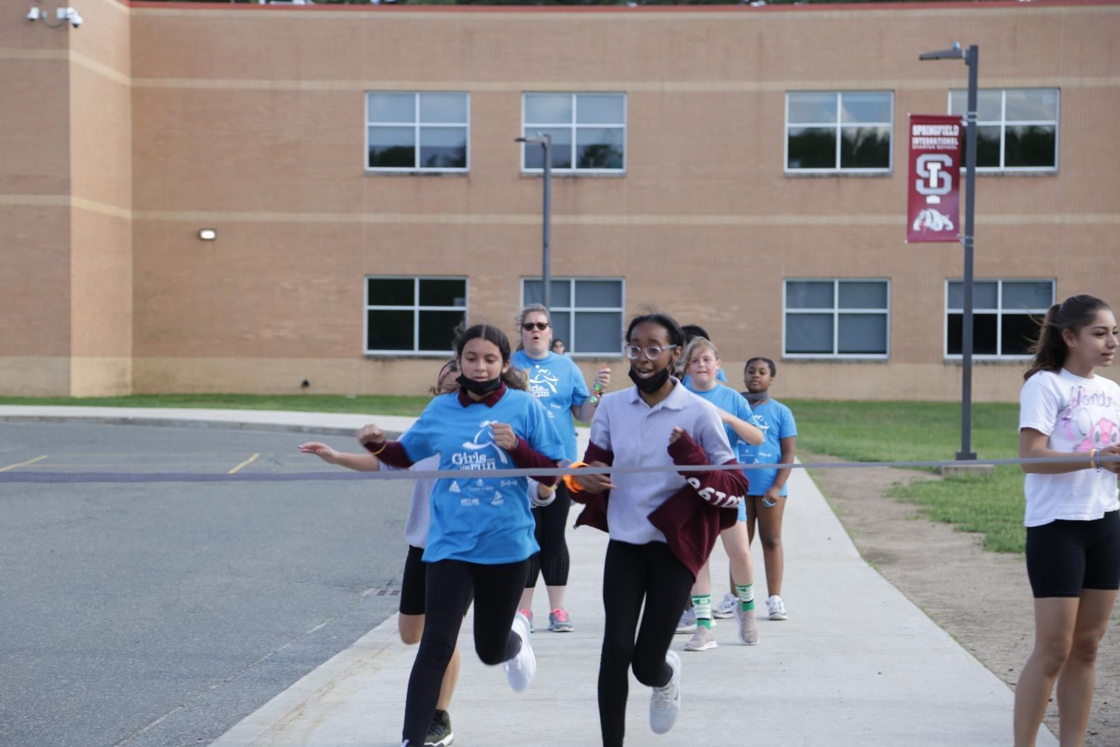 A group of people are running down a sidewalk in front of a building that has the letter s on it