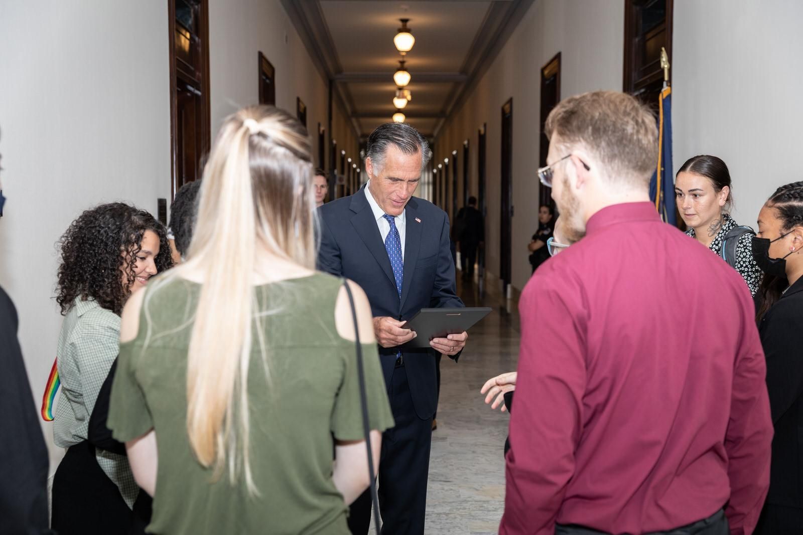 A man in a suit and tie is talking to a group of people in a hallway
