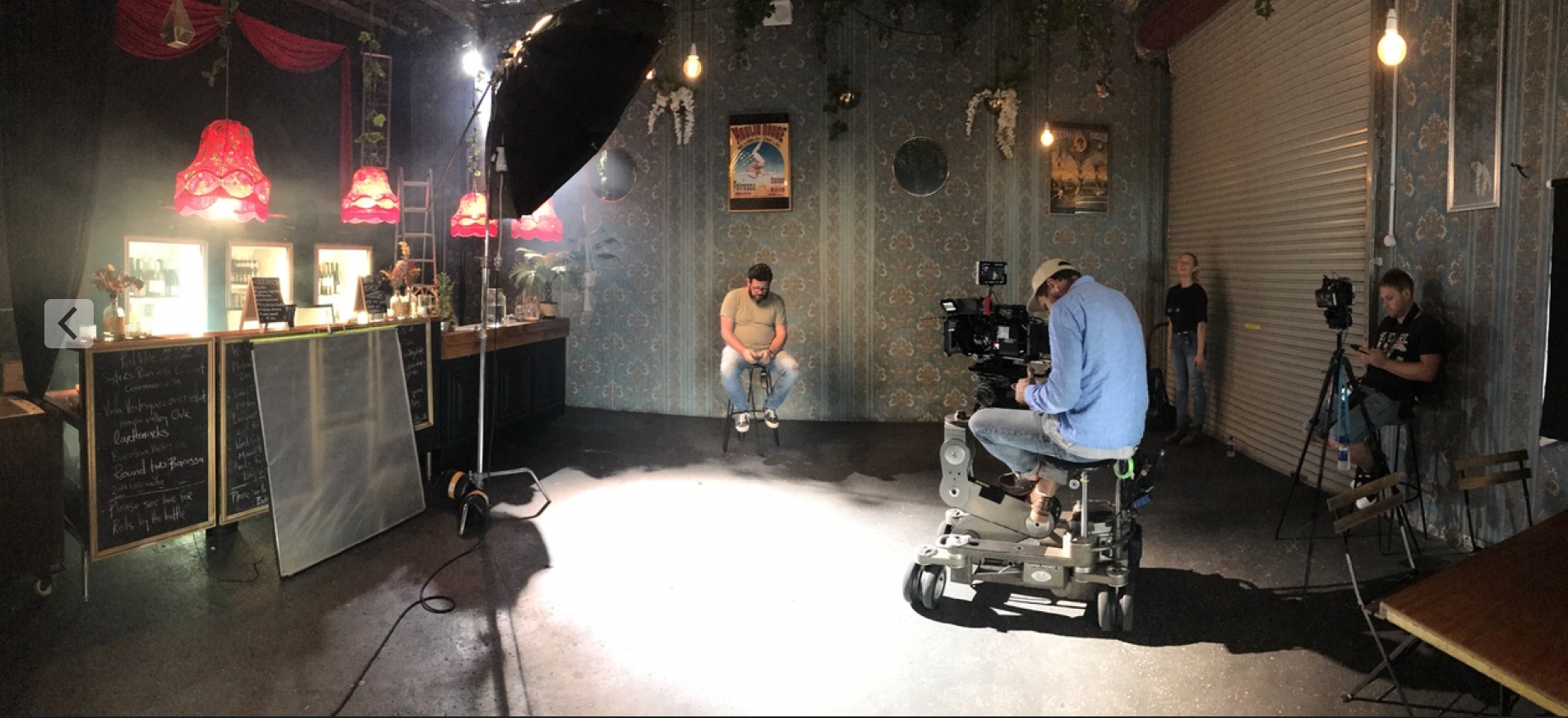 A man is sitting on a stool in front of a camera in a dark room.