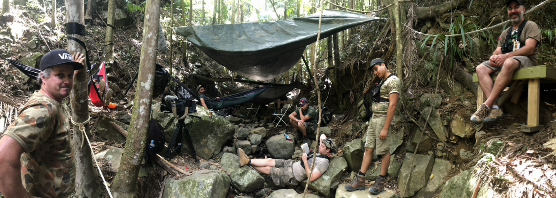 A group of people are standing around a hammock in the woods.