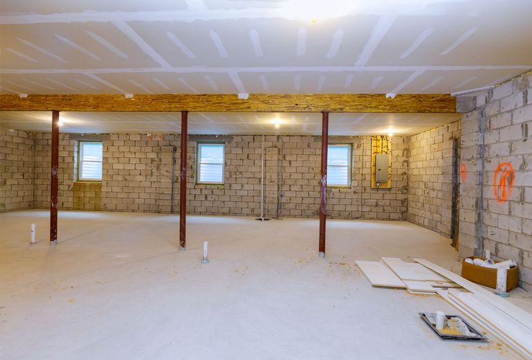 Basement under construction, exposed brick walls, drywall on ceiling, support beams, unfinished floor.