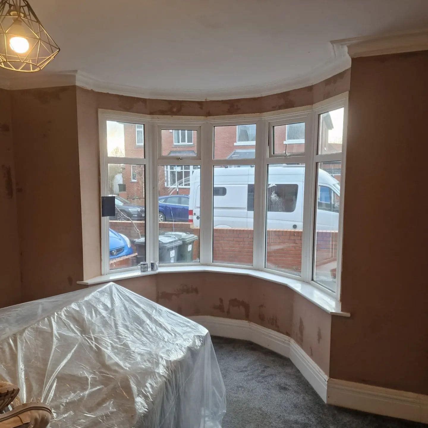 Room with a large bay window, freshly plastered walls, and covered furniture.