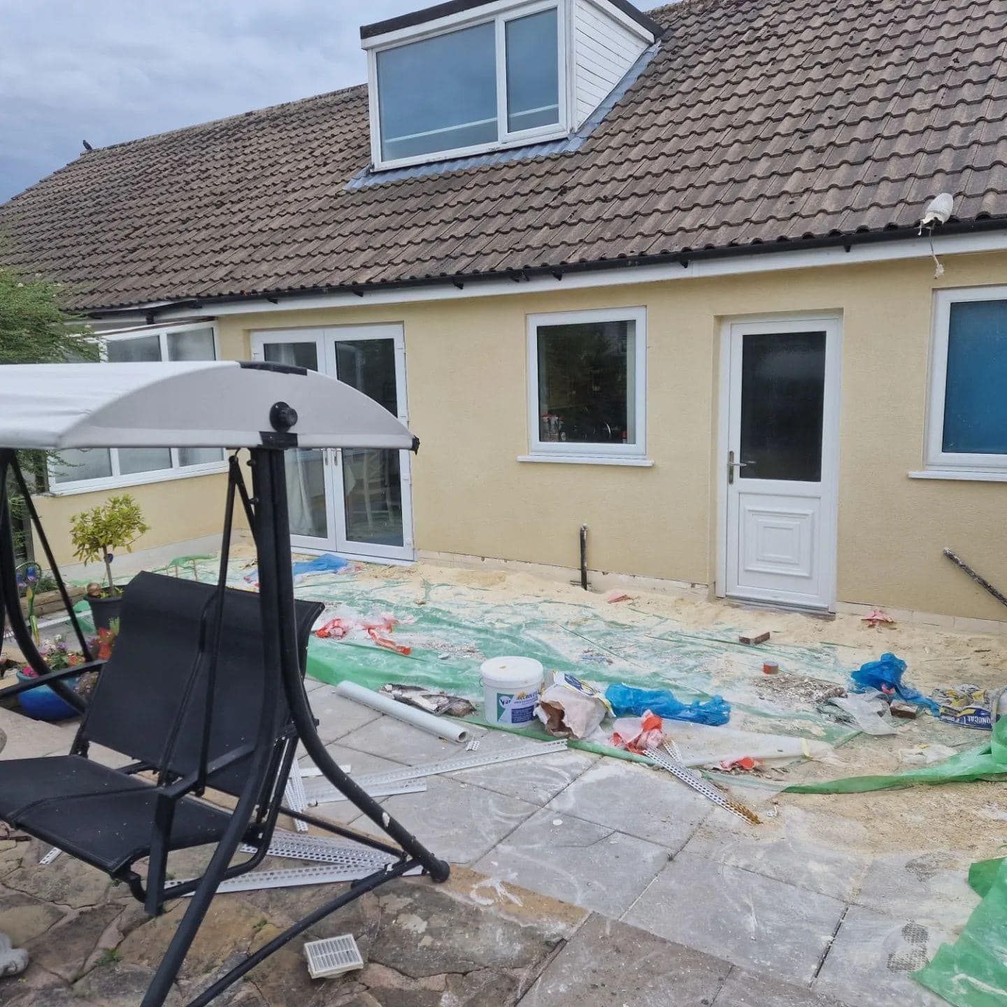 Backyard renovation with swing chair, debris, yellow stucco wall, and white doors.