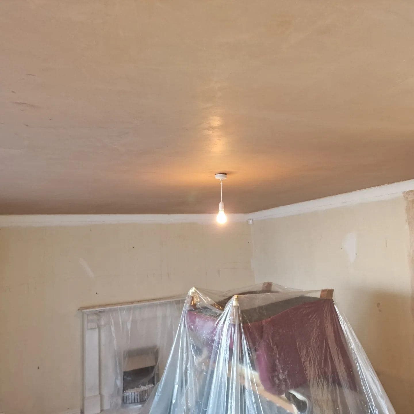 Room with freshly plastered ceiling; fireplace and furniture covered in plastic sheeting.