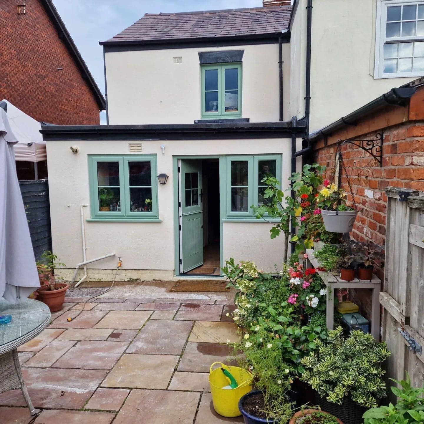 Small cottage with cream walls, green windows, and a brick patio with plants.