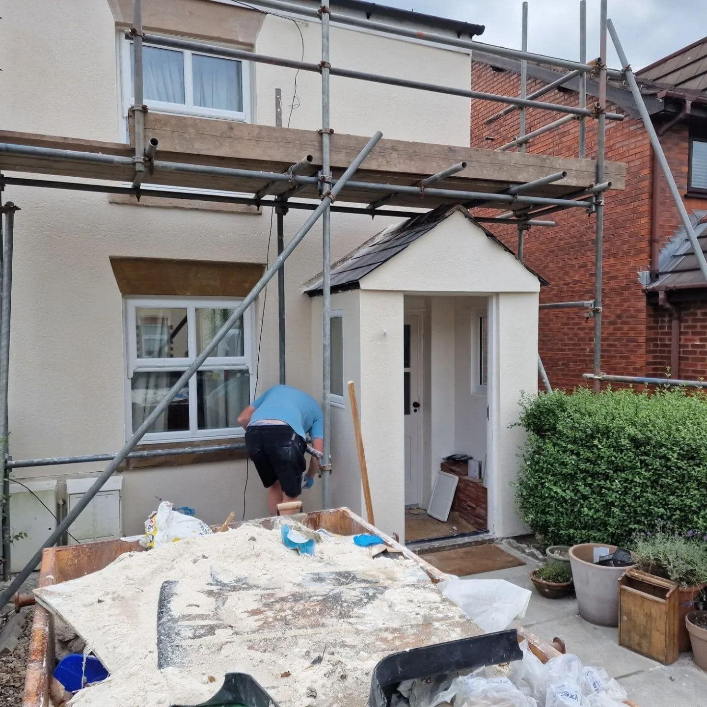 Man works on house exterior with scaffolding, debris in foreground. Beige stucco, white trim.