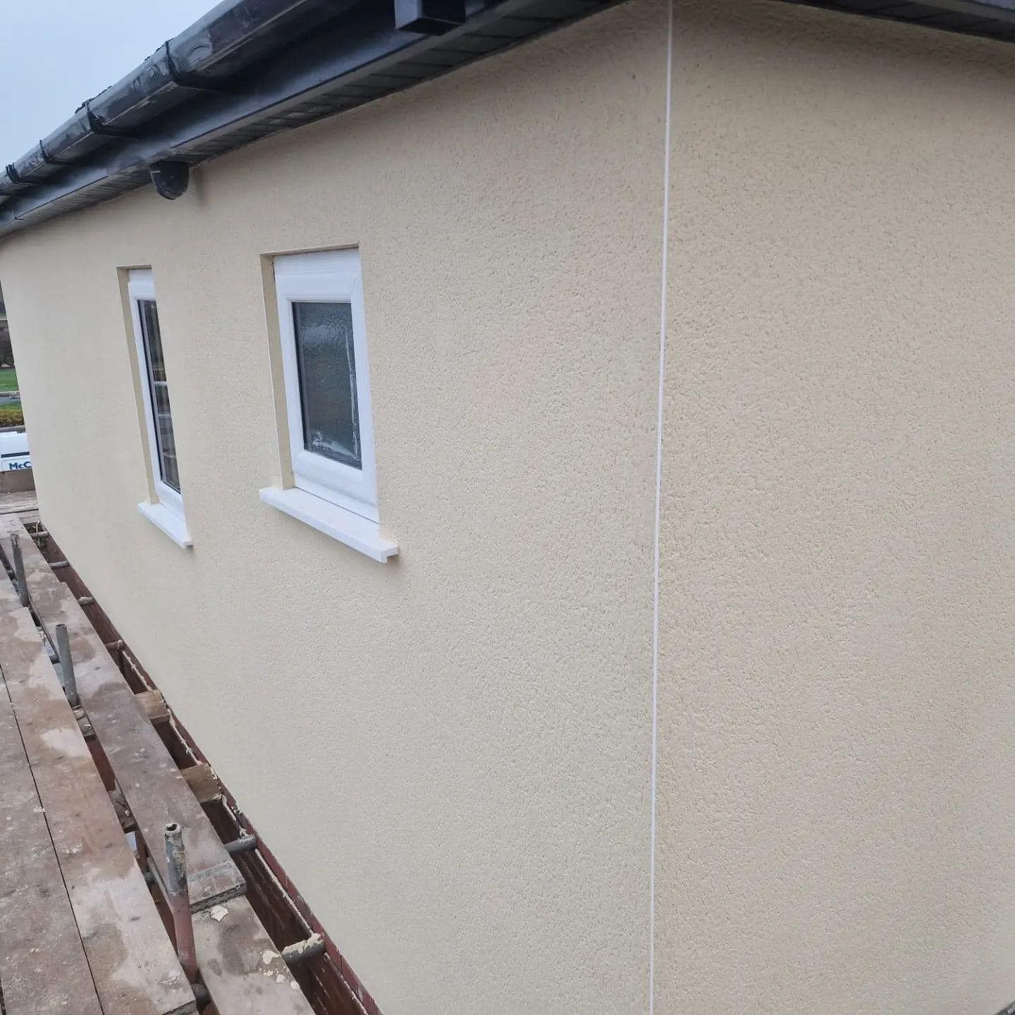 Beige stucco building with two white-framed windows, corner detail, and dark roof.