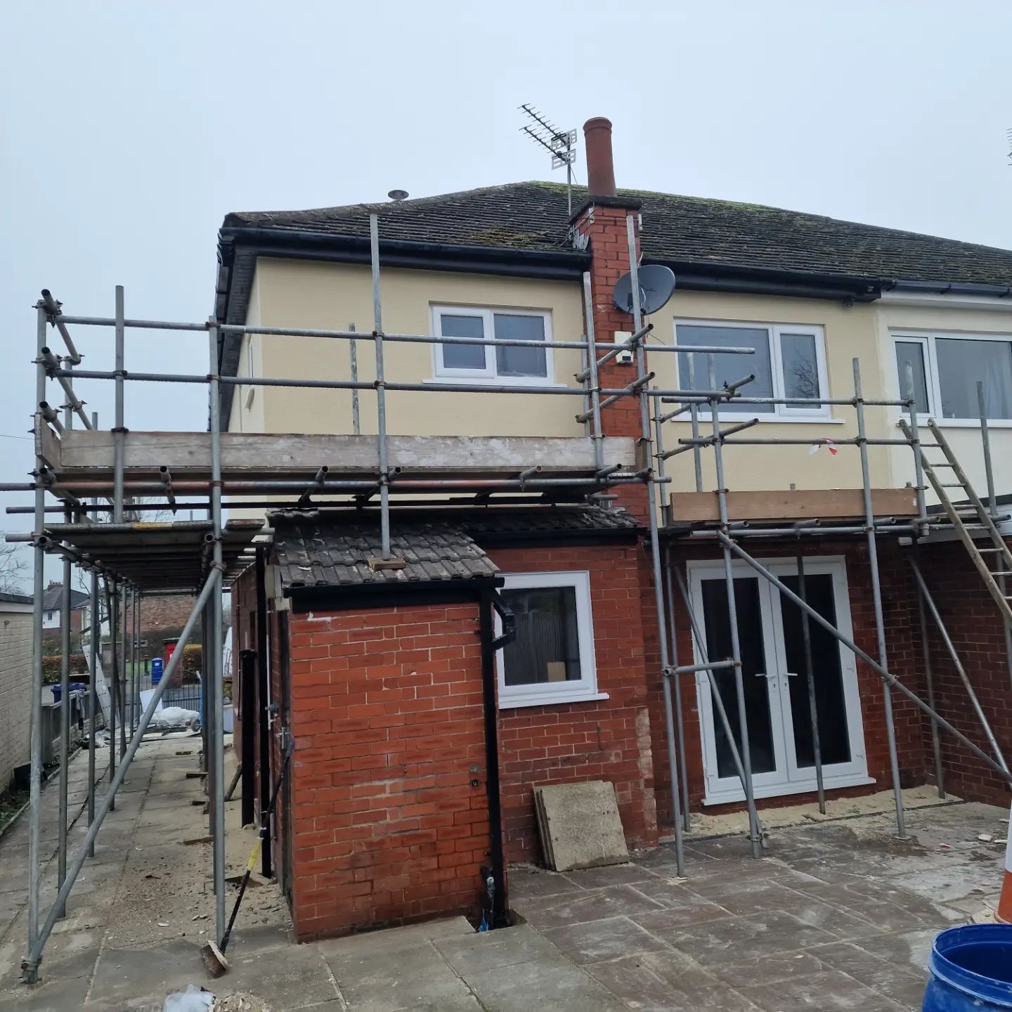 Scaffolding erected on the back of a house with a brick extension, cloudy sky.