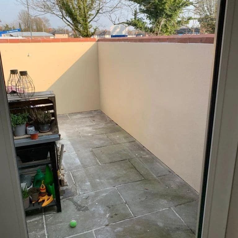 Small patio with gray stone tiles, cream walls, and wooden furniture. Sunlight.