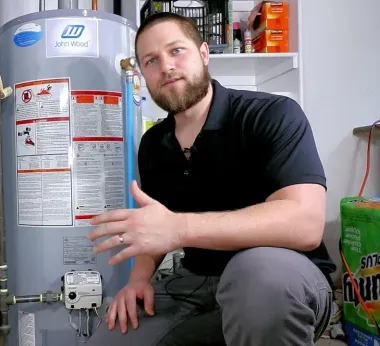 Man kneeling by a water heater, pointing to a valve. He's in a utility room.