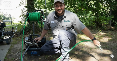Man in green shirt kneels by a power washer, holding a hose in outdoor setting.