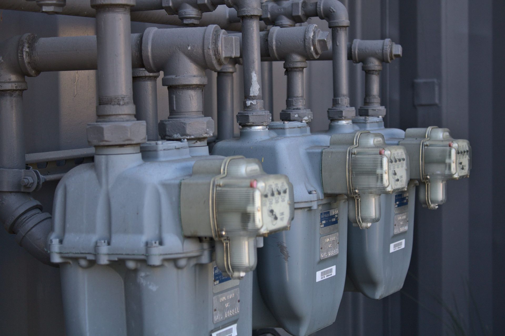 Close-up of gray gas meters with pipes against a dark gray building.