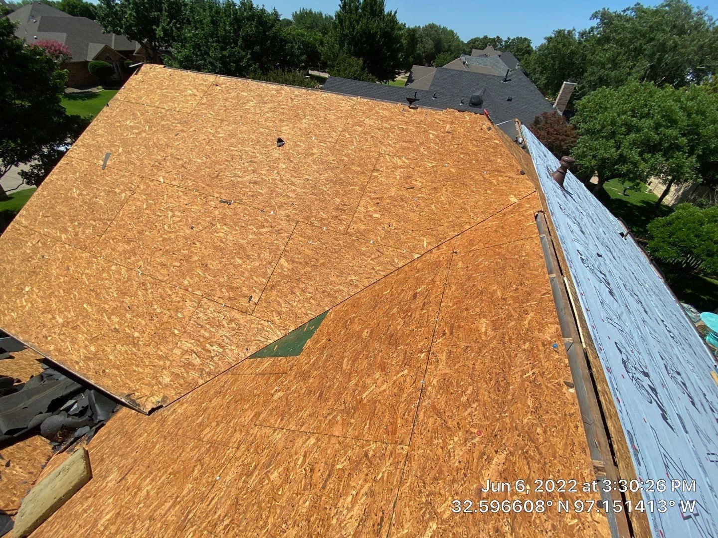 Rooftop with exposed OSB sheeting, some underlayment, and surrounding trees in a residential area.