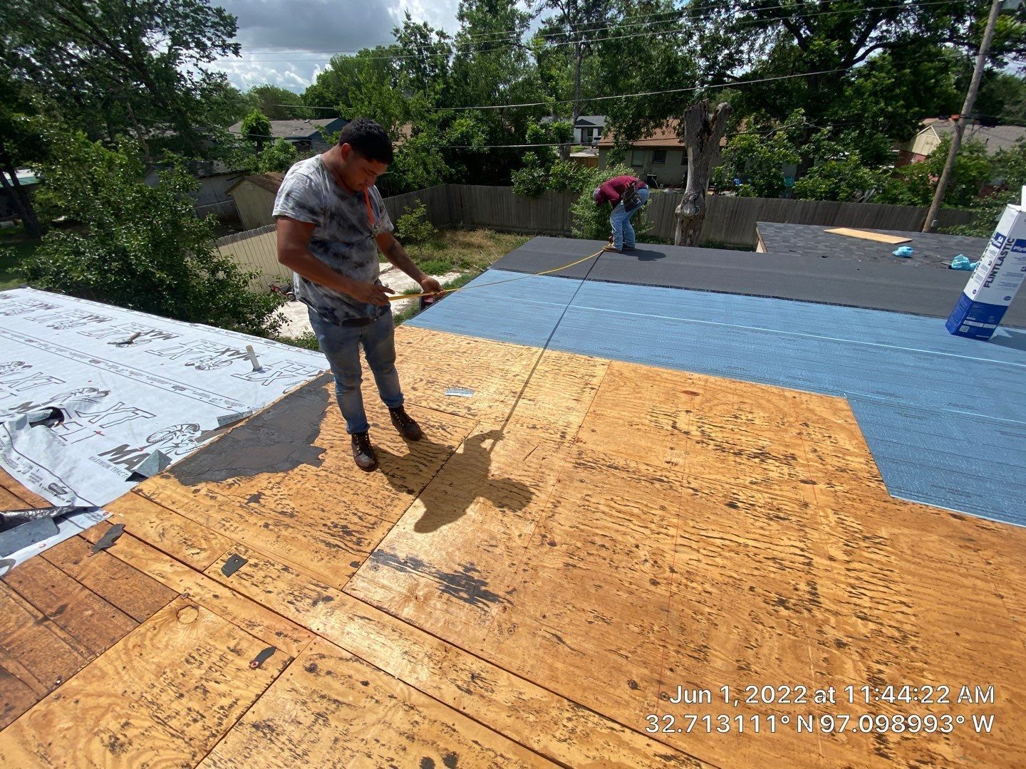 Two workers on a rooftop, one applying a product. Wooden roof partially covered with blue underlayment, trees in background.