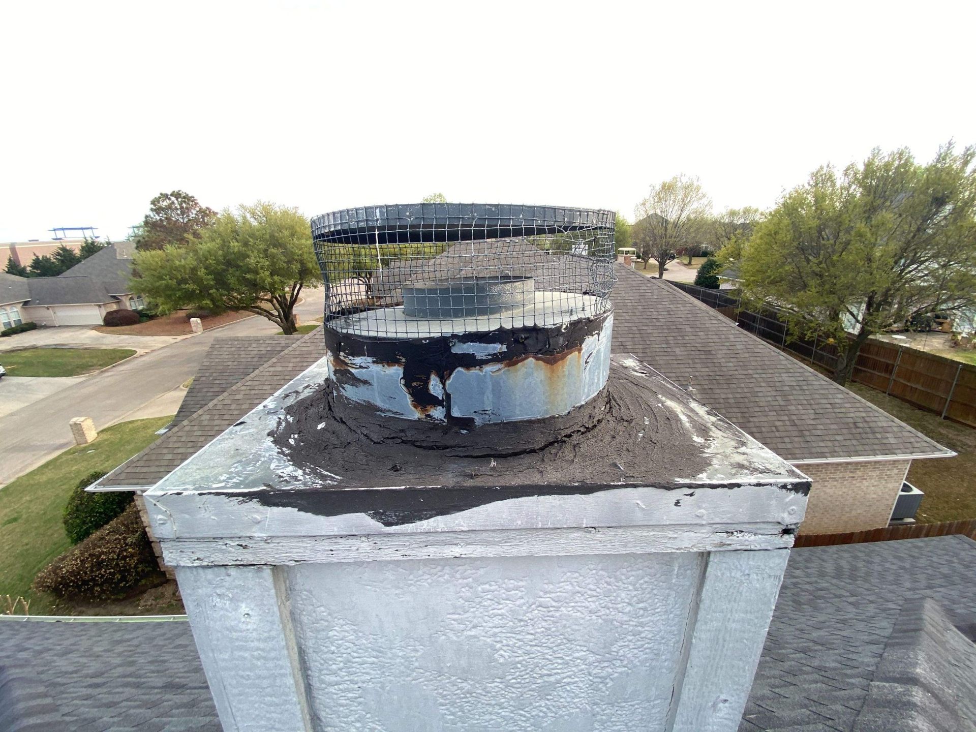Chimney with a damaged metal cap and mesh screen on a rooftop in a suburban neighborhood.