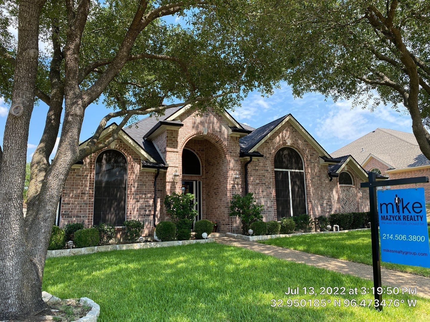 A brick house for sale, with a sign in the front yard, framed by trees under a blue sky.
