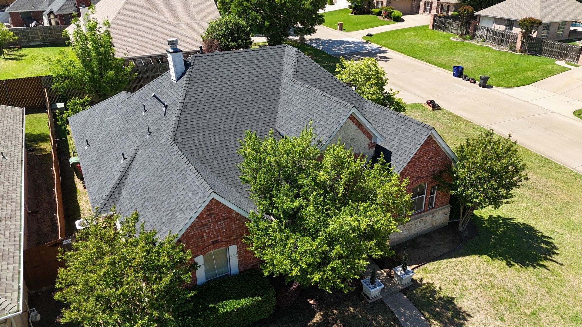 An aerial view of a brick home with a dark gray roof, surrounded by green trees and grass.