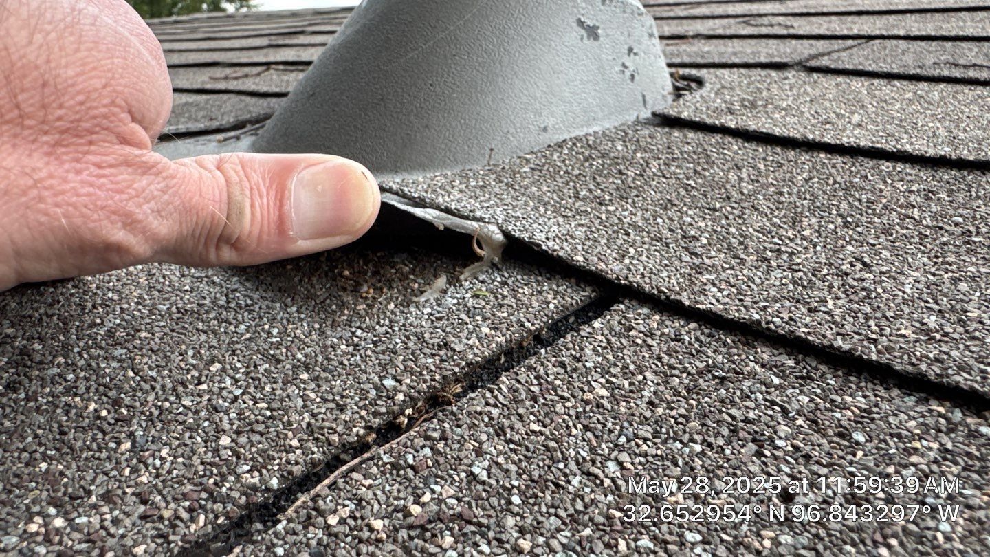 Hand lifting a weathered asphalt shingle near a roof vent, possibly inspecting for damage.