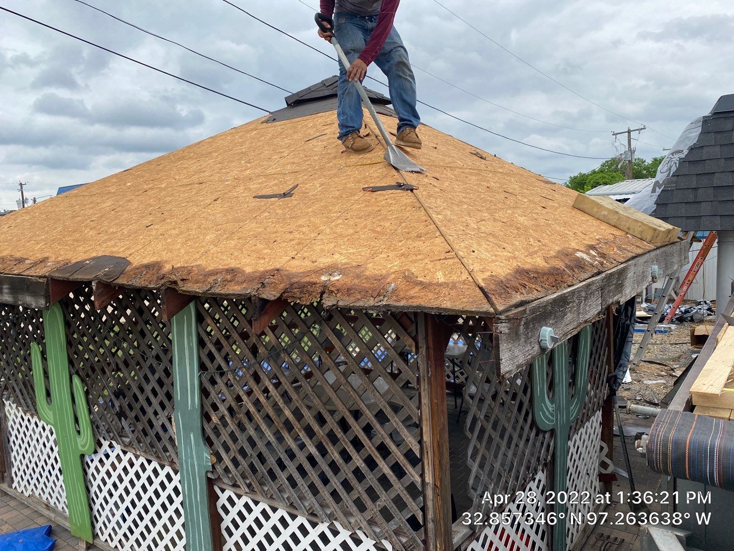 Person on rooftop removes shingles from a gazebo with a shovel. Cloudy sky in the background.
