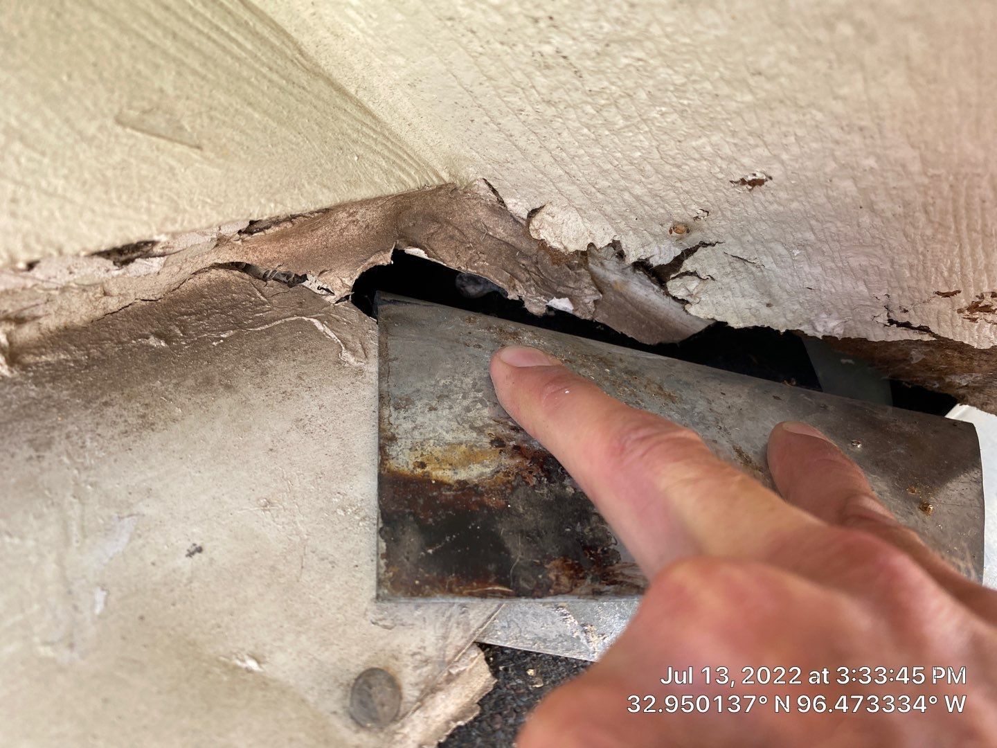 Finger pointing at a rusty metal plate inside a hole in a deteriorated wall.