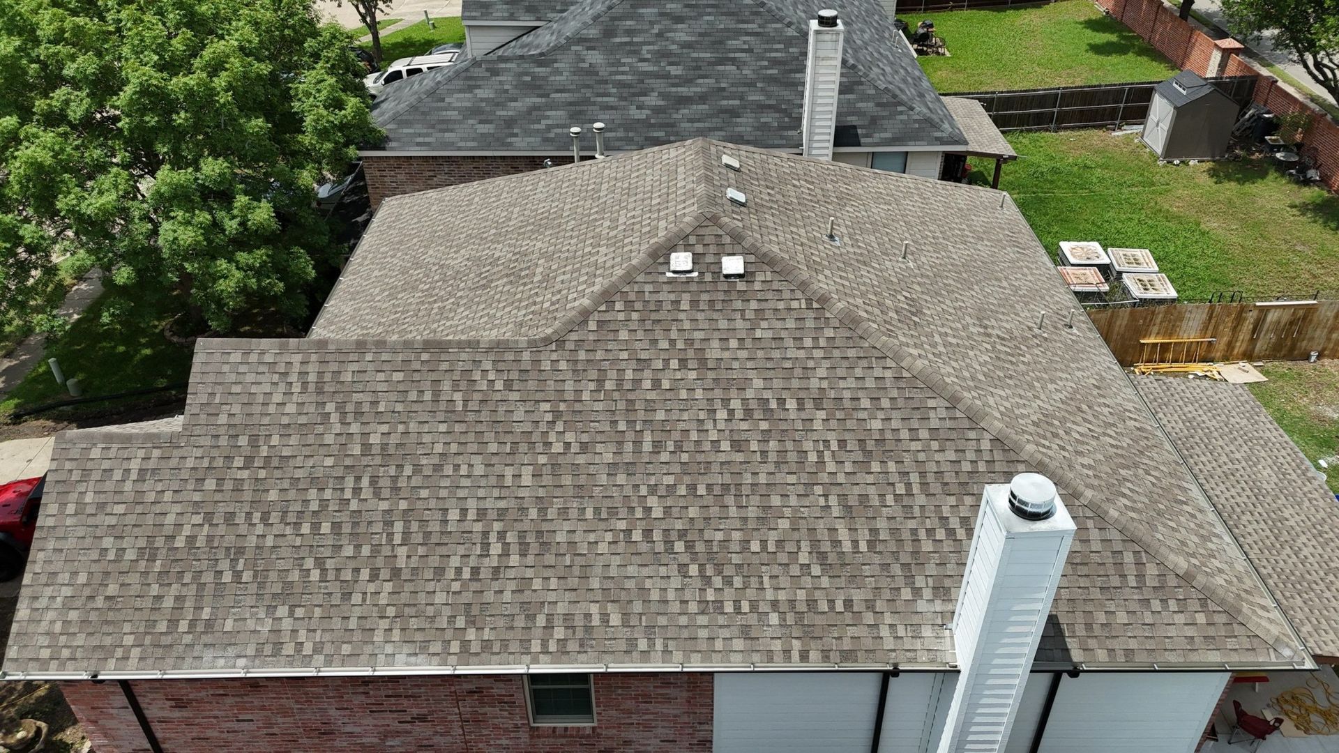 Aerial view of a brown shingled roof with a brick and white house. A chimney is visible.
