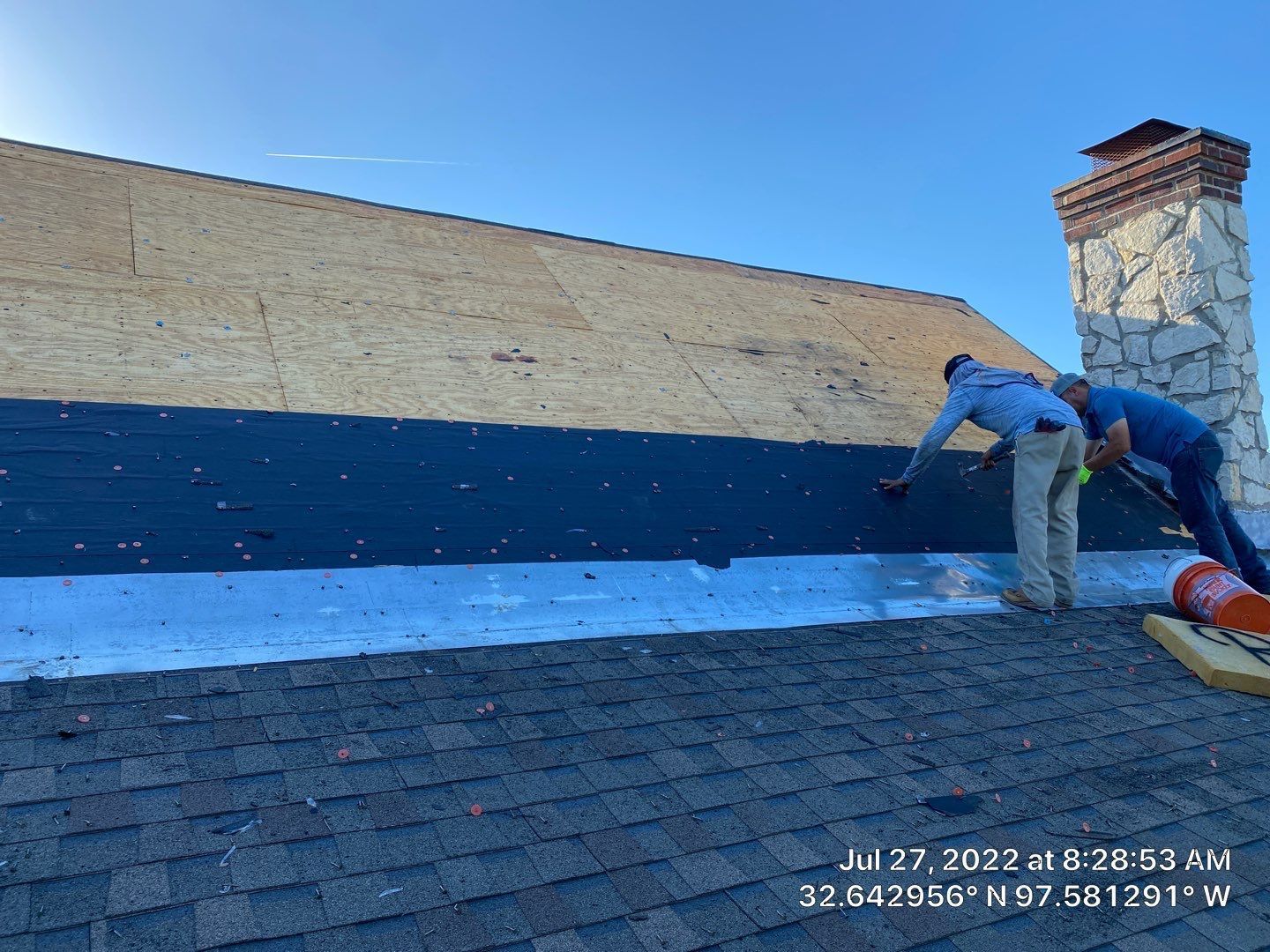 Two roofers installing black roofing material on a house roof near a chimney.