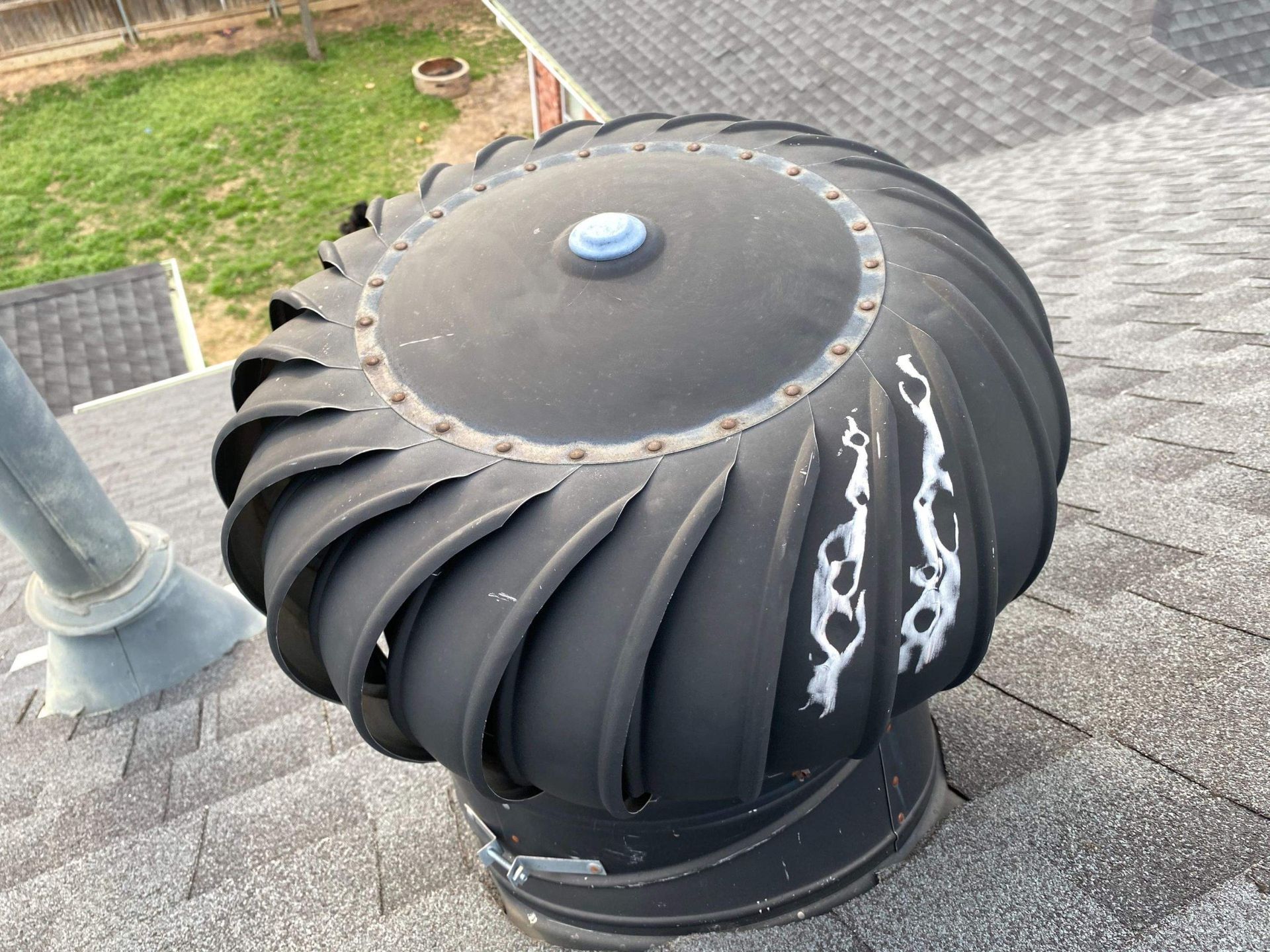 Black turbine roof vent on a shingled roof, next to a metal pipe.