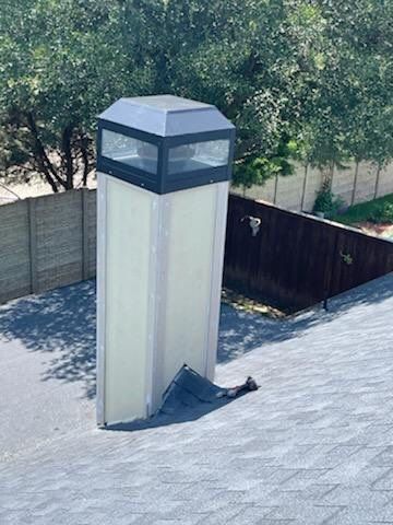 Tall rectangular skylight on a gray shingle roof, surrounded by fences and trees.