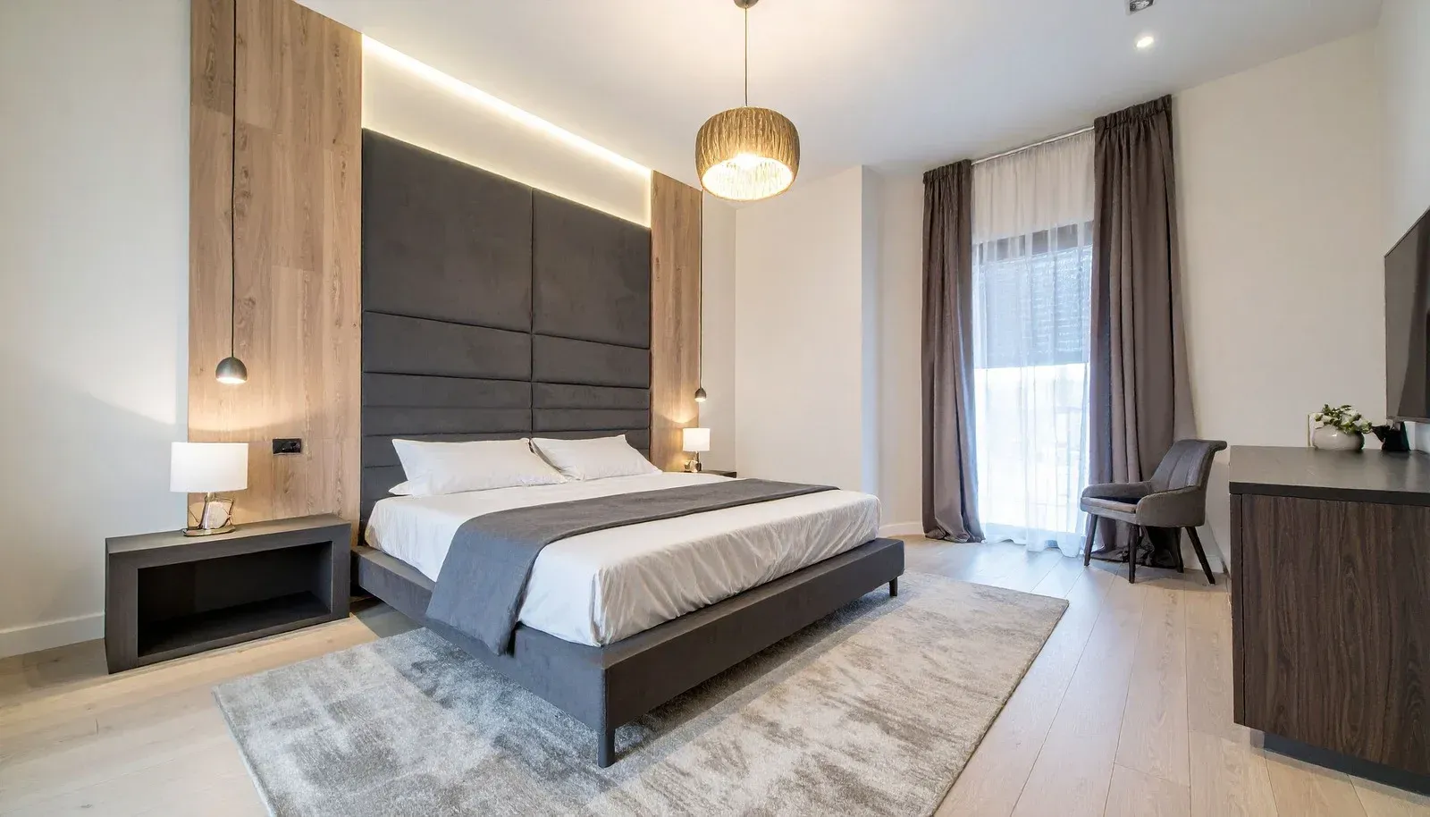 Modern hotel bedroom with large bed, gray headboard, wood paneling, and a rug.
