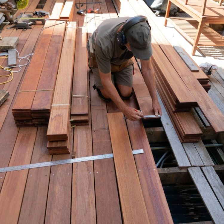 Man kneeling, measuring wood planks for a deck with stacked boards in the sunlight.