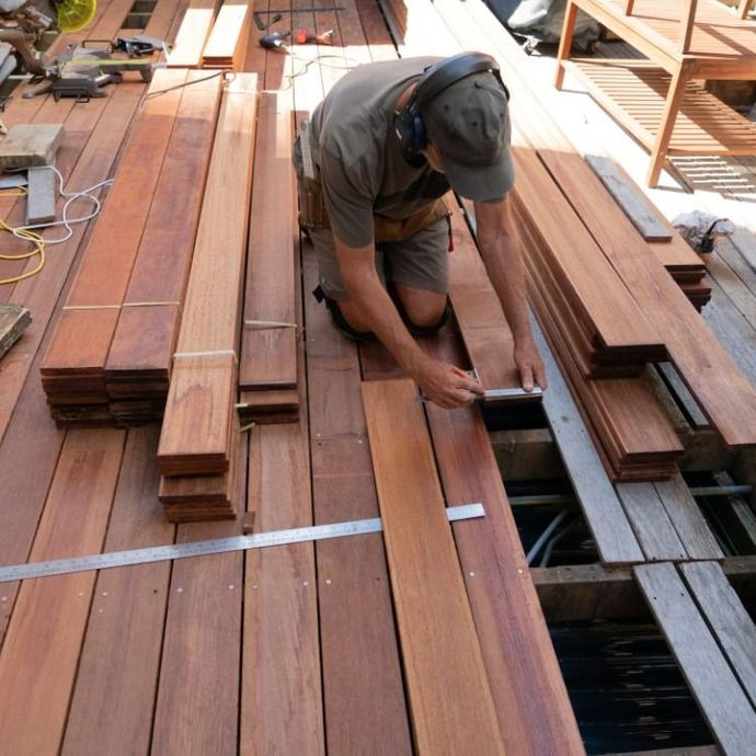 Man kneeling, measuring wood planks for a deck with stacked boards in the sunlight.