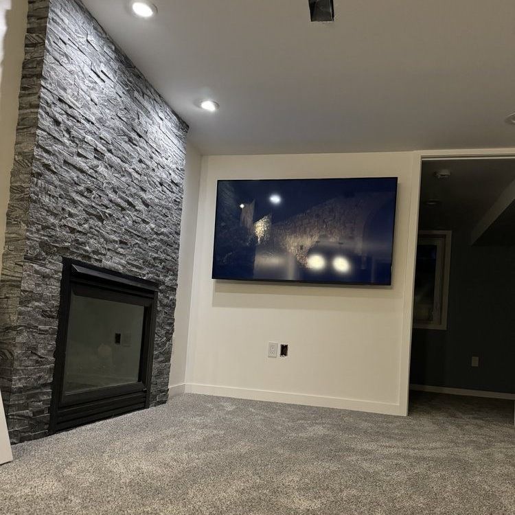 A modern living room with a stone fireplace, a wall-mounted TV, and gray carpet.