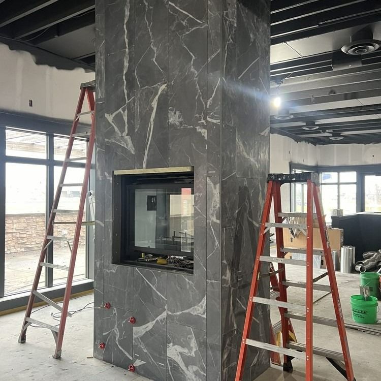 Fireplace under construction, dark gray marble tile, two ladders, unfinished interior.