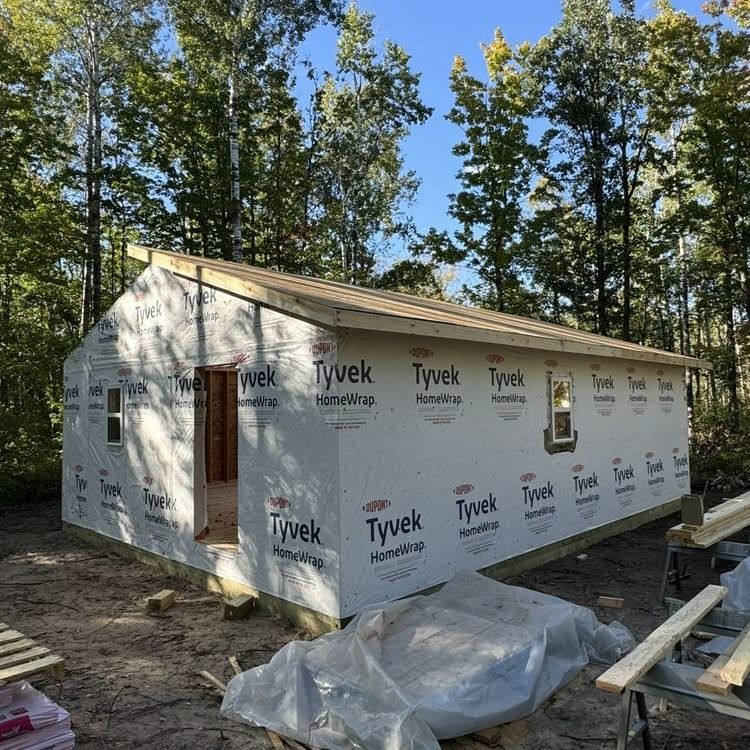 A small building under construction, wrapped in Tyvek, in a wooded area.