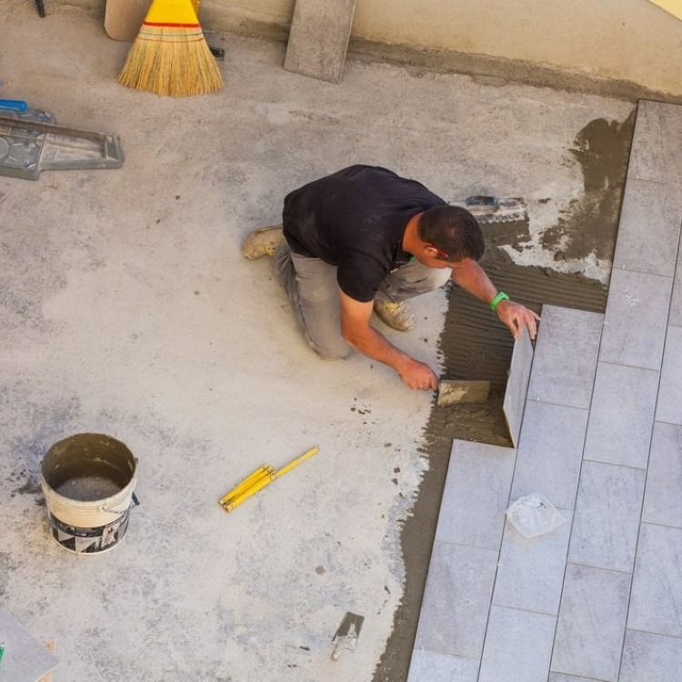 Person laying tiles on a concrete surface, using tools and mortar.
