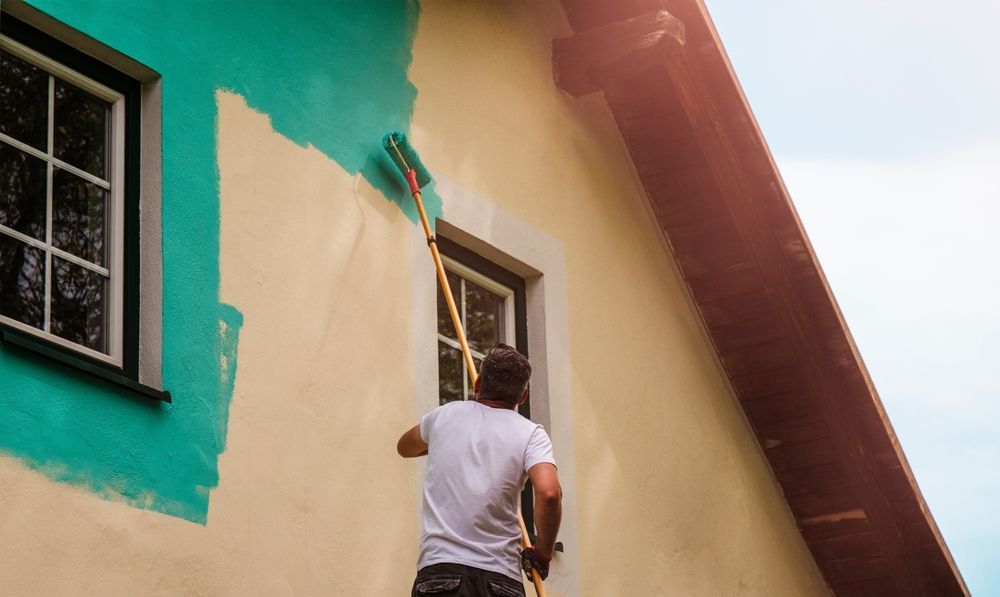 Man painting exterior wall teal with a roller; house facade.