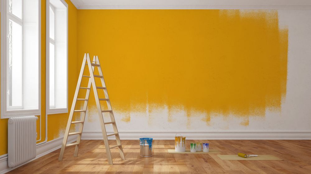 Interior room being painted yellow, ladder, paint cans, brush, partially painted wall, wood floor, white trim.